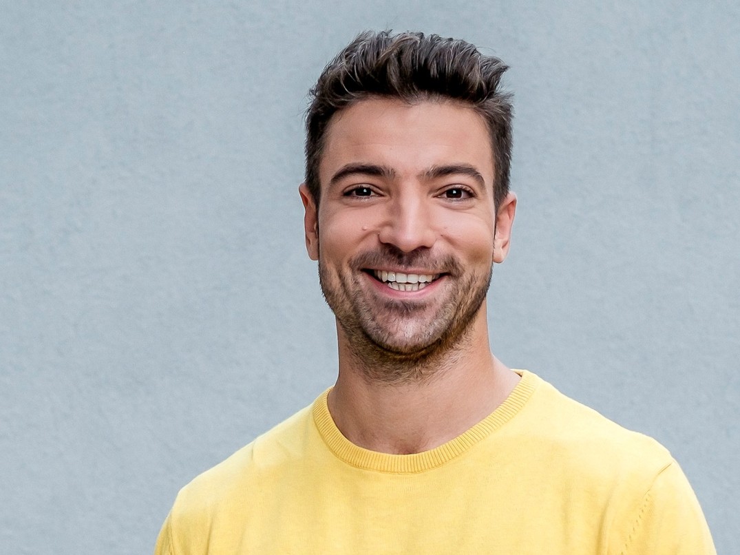 Ramon Babazadeh Biografie: Porträtfoto des deutschen TV-Moderators in gelbem Pullover mit einem offenen, sympathischen Lächeln vor grauem Hintergrund.